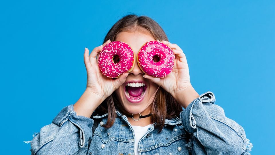 Woman holds two brightly colored donuts over her eyes. Her mouth is open wide in a playful expressions.